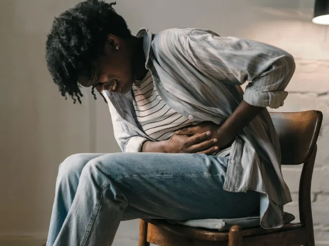 an individual, seated on a wooden chair, clutching their stomach in discomfort