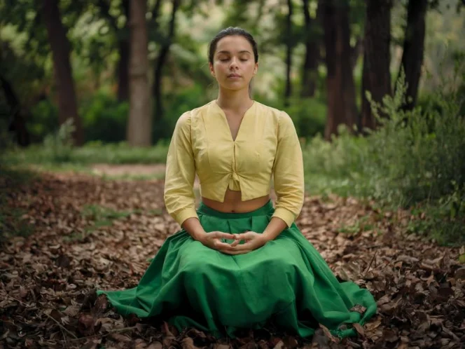 A photo of a woman sitting in a serene forest. She is wearing a yellow blouse and a green skirt
