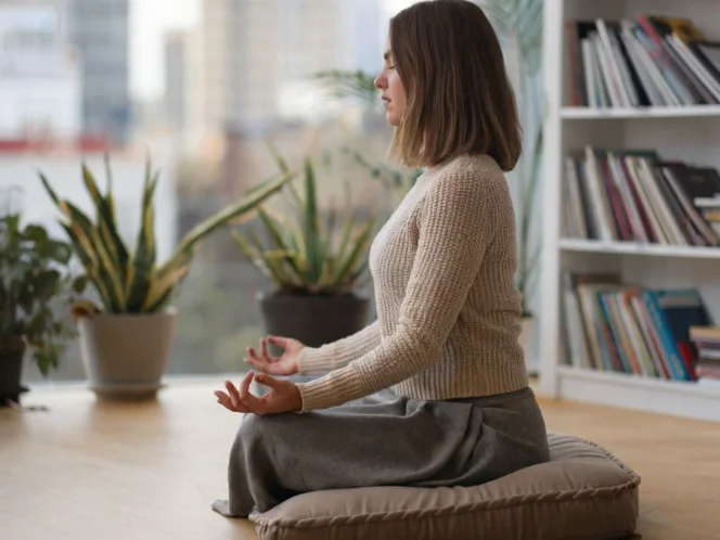 A photo of a woman with shoulder-length brown hair meditating on a cushion on a wooden floor